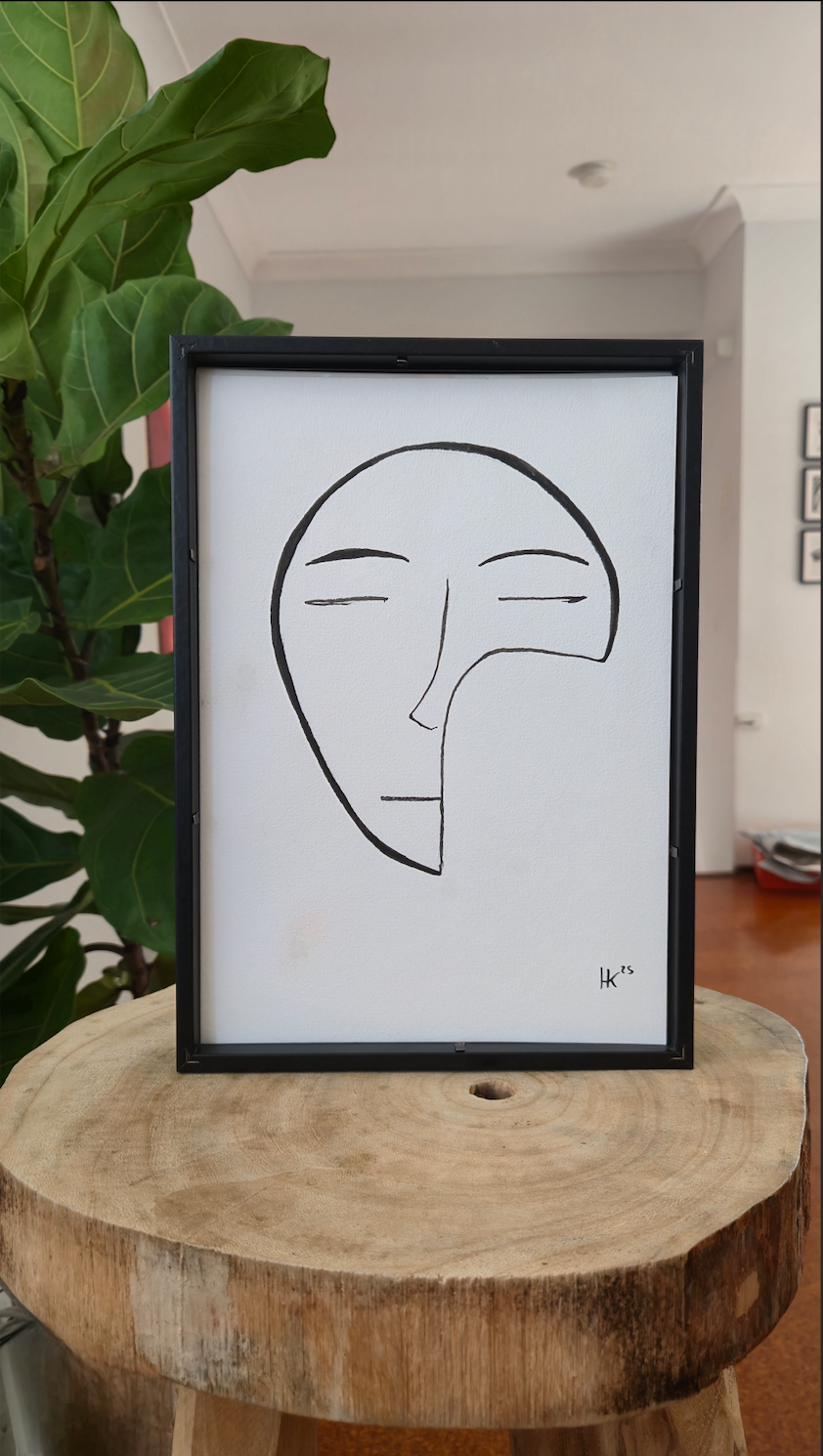 Framed abstract face drawing on a wooden stool with a plant in the background