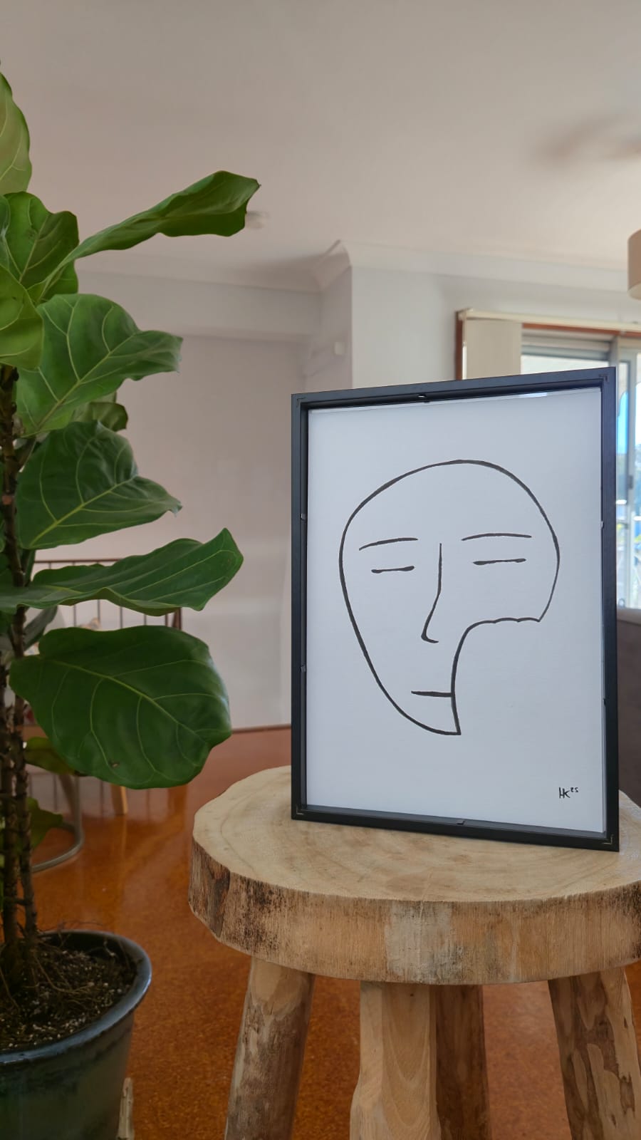 Framed abstract line art of a face on a wooden stool with a plant in the background