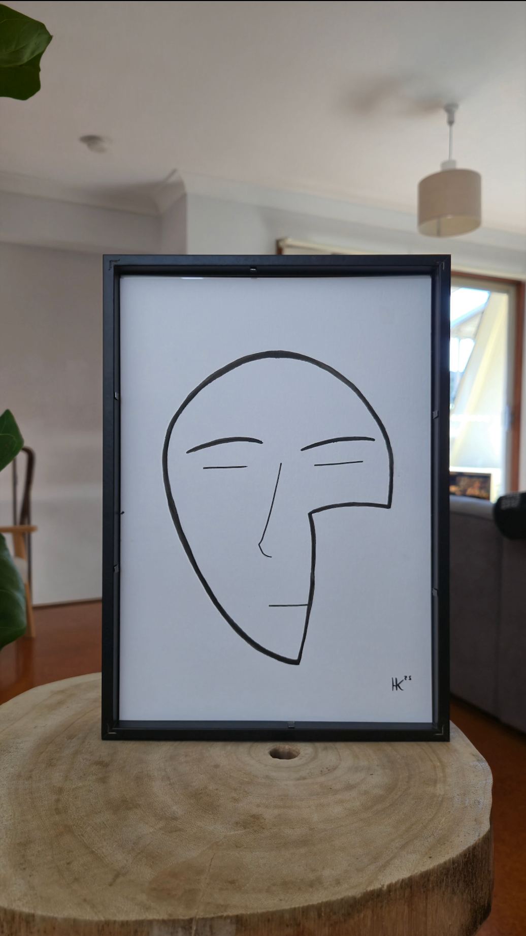 Framed black and white line drawing of a face on a wooden surface with a room background.